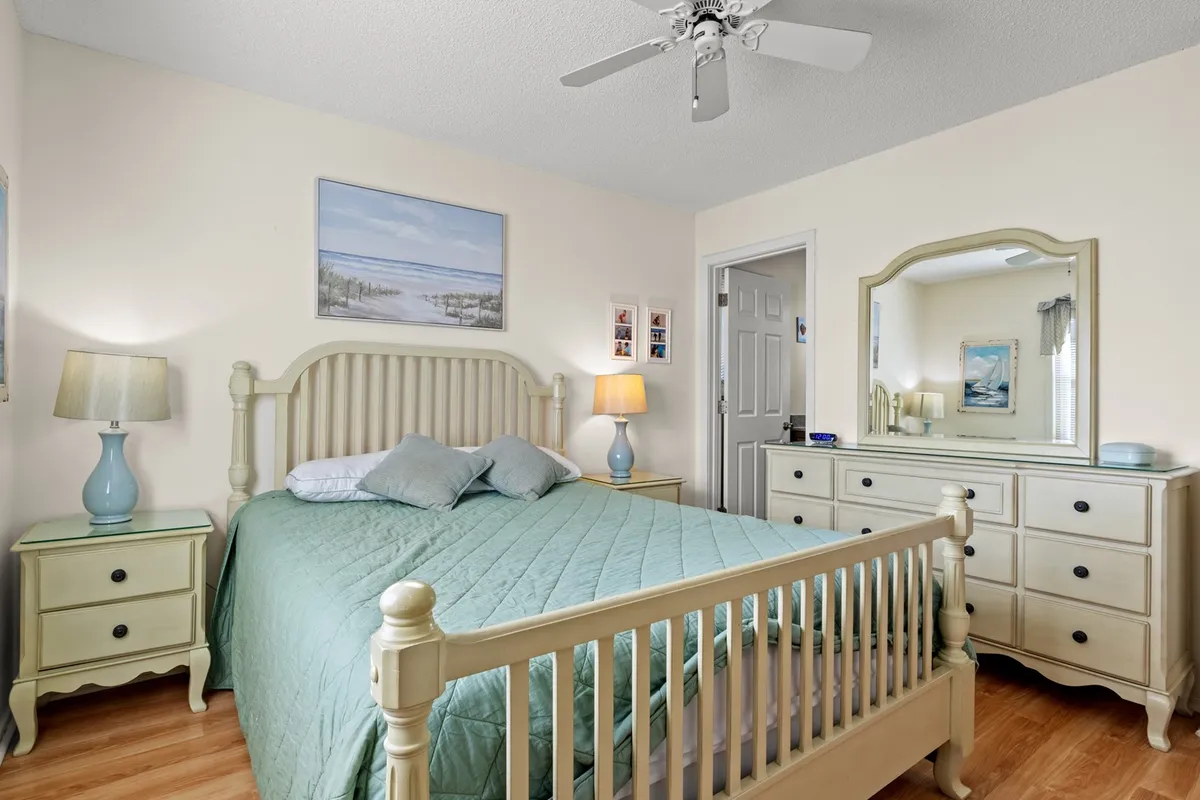 A serene and cozy bedroom featuring a queen bed dressed in a light green quilt, a nightstand with a warm lamp, a dressing table with a mirror, and framed artwork on the wall. Sunlight gently streams through the window, bathing the room in a calming glow.