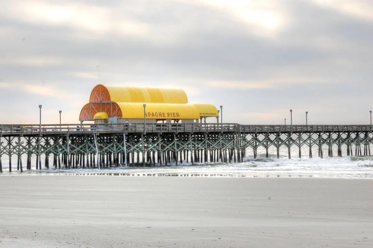 A scenic view of Apache Pier with its colorful yellow roof and expansive wooden walkway extending into the ocean. The setting is ideal for couples or friends to stroll, watch sunsets, or enjoy the vibrant coastal atmosphere.