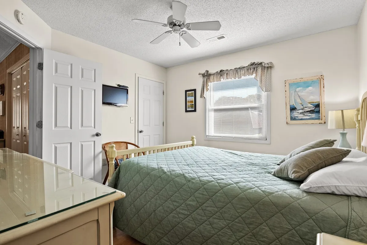 A calm, inviting bedroom with a comfortable bed covered in a green quilt, a window allowing natural light to pour in, decorative artwork, and a small television. The space feels intimate and perfect for relaxation or unwinding with a book or show.