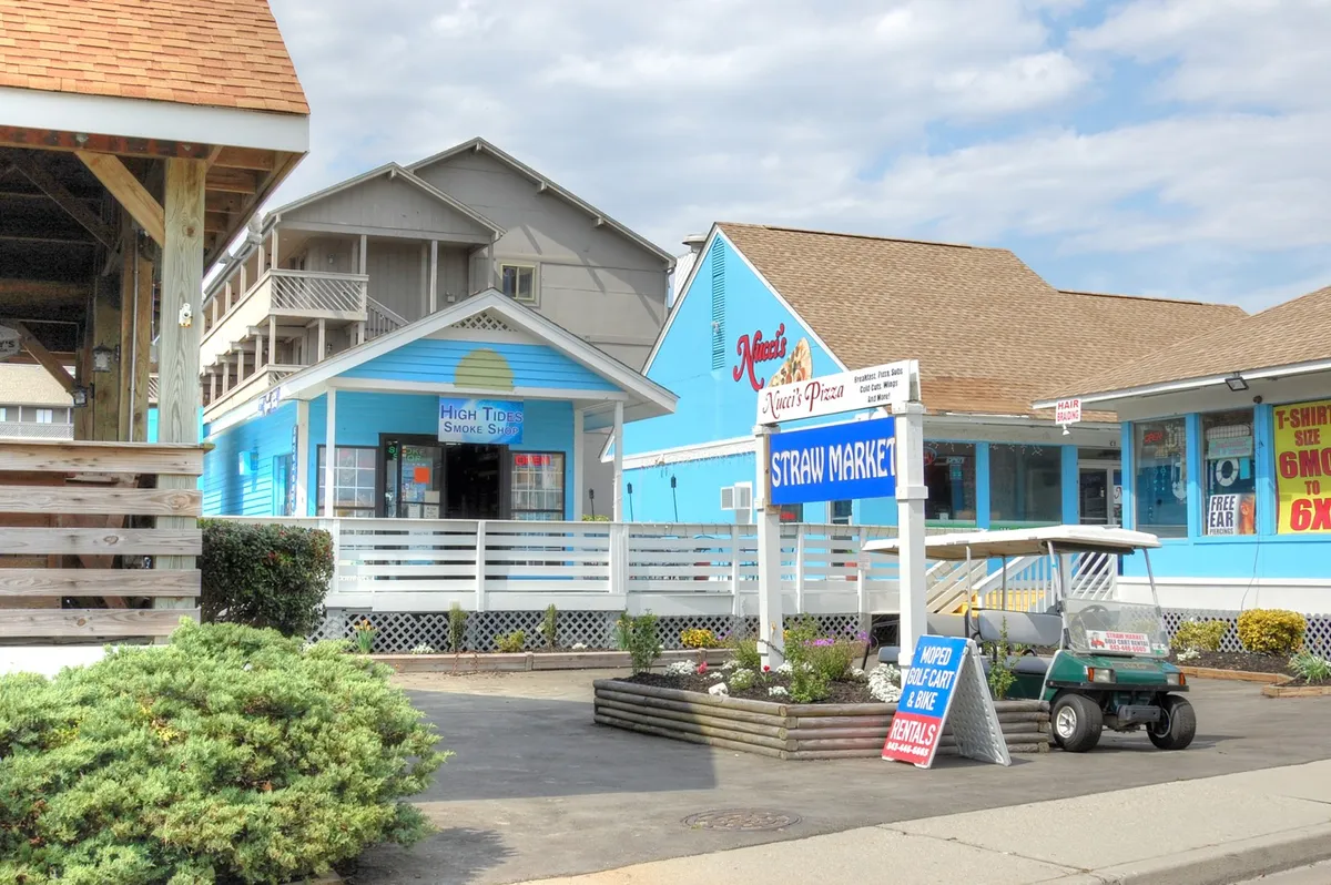 A lively cluster of colorful storefronts, including signage for Straw Market and a pizza place, with a golf cart in the foreground. The scene is bustling and friendly, illustrating local shopping and dining opportunities.