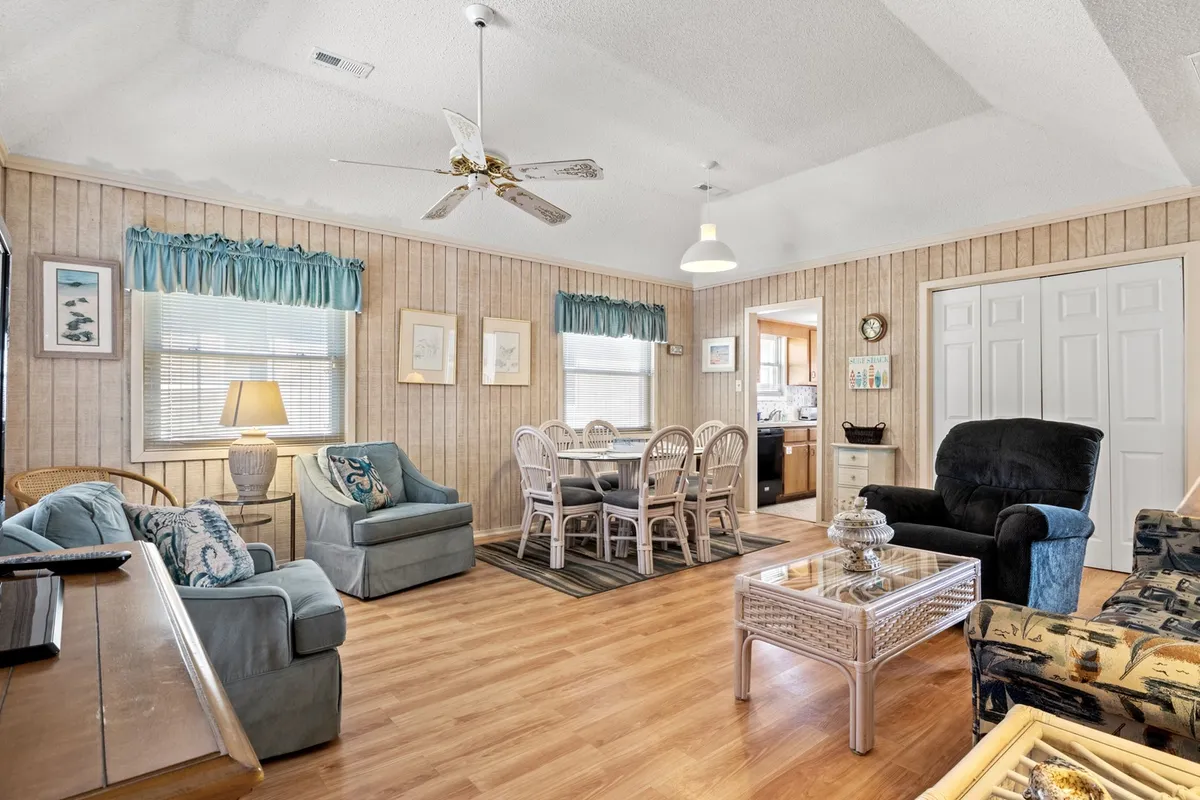 This image showcases a welcoming open-plan living and dining area. Soft natural light pours in through large windows, illuminating a plush sofa with decorative pillows, a spacious dining table with several chairs, and a central coffee table. The ceiling fan and tasteful décor contribute to a relaxed, homey atmosphere ideal for socializing or winding down.
