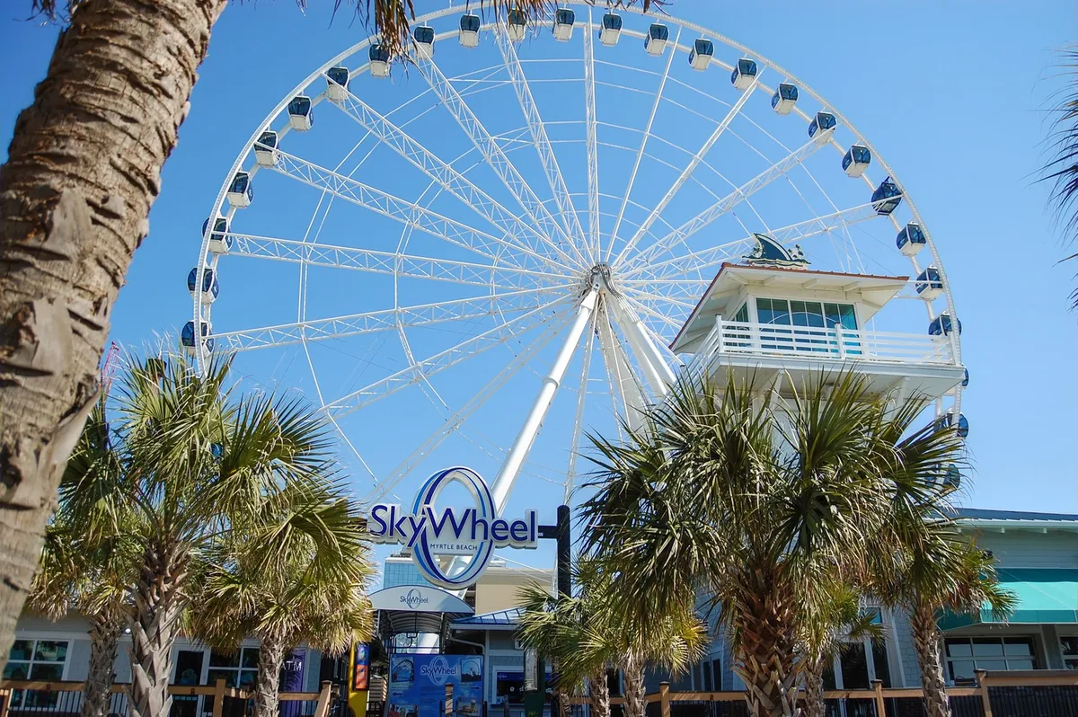 A vibrant view of the SkyWheel surrounded by palm trees, with a lively ticket booth in the foreground. The scene is bright and cheerful, capturing the joy and excitement of this iconic Myrtle Beach attraction.