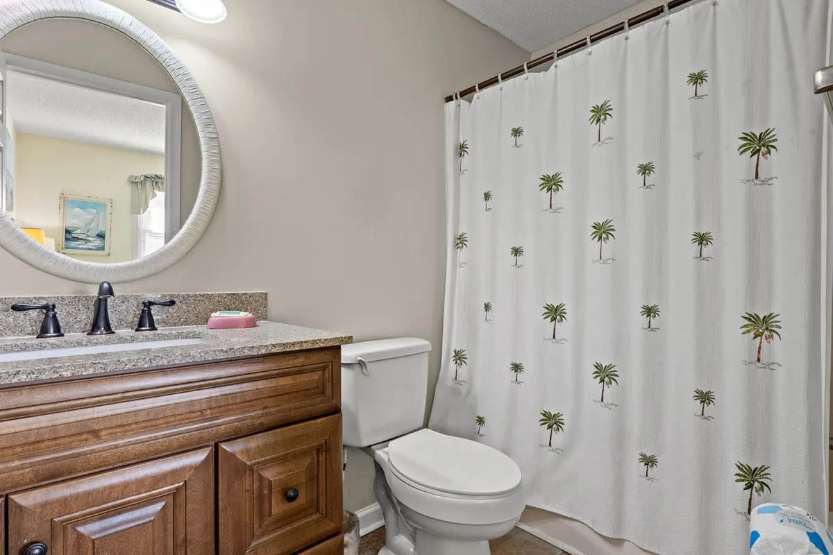 A tranquil bathroom with a well-lit vanity and sink, a palm tree-themed shower curtain adding a touch of fun, and a round mirror above the countertop. The scene is fresh, bright, and inviting, ideal for morning routines or a relaxing bath after a busy day.