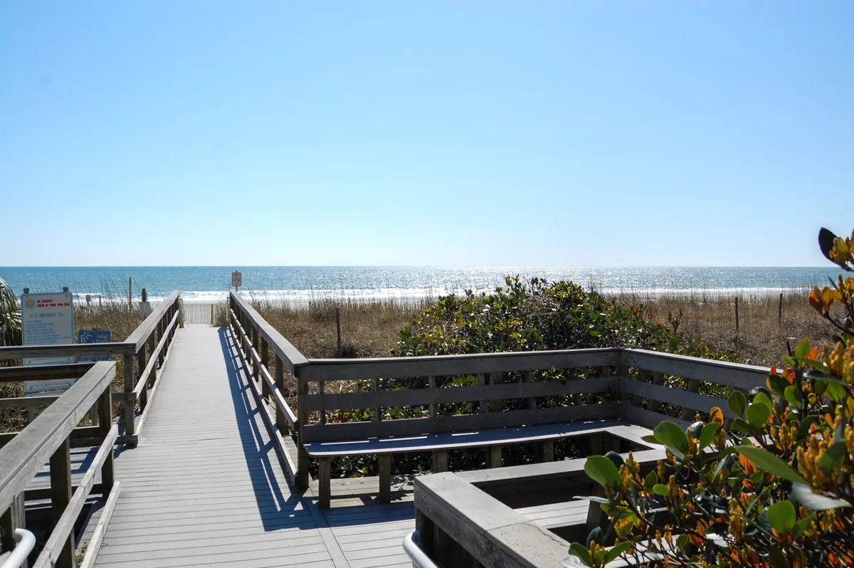 A wooden boardwalk stretches out towards the ocean, bordered by dune vegetation and offering expansive views of the water. The image exudes a tranquil, natural vibe, inviting guests to take peaceful walks along the beach.