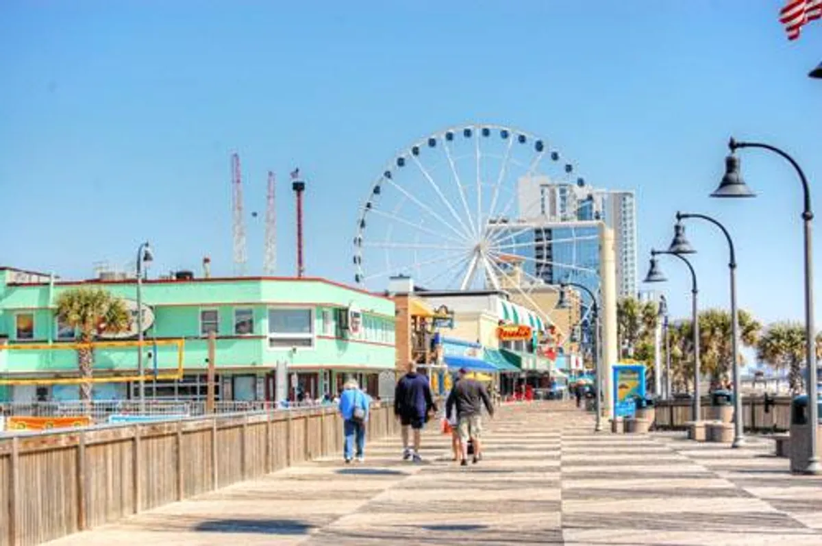 A vibrant boardwalk scene at nearby Broadway at the Beach, featuring a colorful Ferris wheel, lively buildings, and people strolling. The atmosphere is energetic and joyful, capturing the wide range of entertainment, dining, and shopping options available to groups and families.