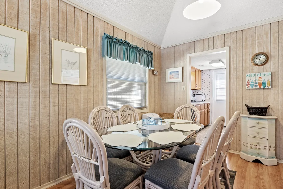 A bright and welcoming dining area with a glass-topped table and woven chairs, set next to a sunlit window dressed with curtains. Decorative wall art and a warm color palette foster a sense of togetherness, making it ideal for shared meals or planning adventures.